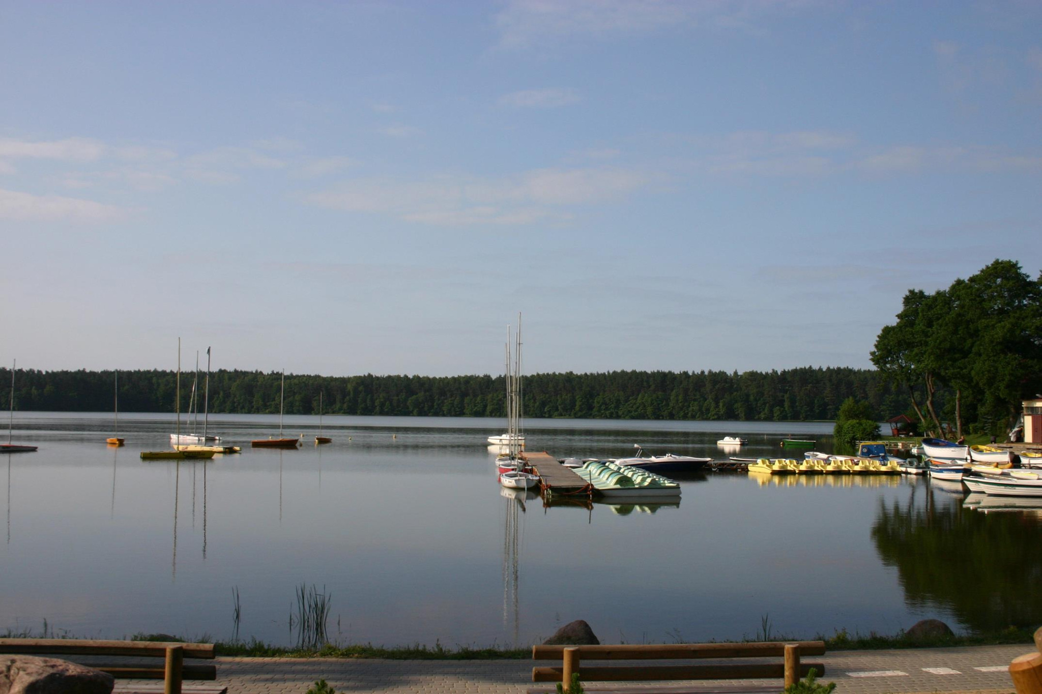 Spokojne jezioro z pomostem, zacumowanymi łódkami żaglowymi i rowerkami wodnymi w słoneczny dzień, widok z perspektywy brzegu z ławkami.