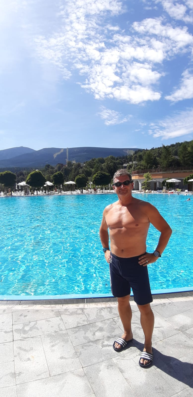 Mężczyzna w kąpielówkach i okularach przeciwsłonecznych stoi na tle turkusowego basenu z widokiem na góry i błękitne niebo z chmurami.