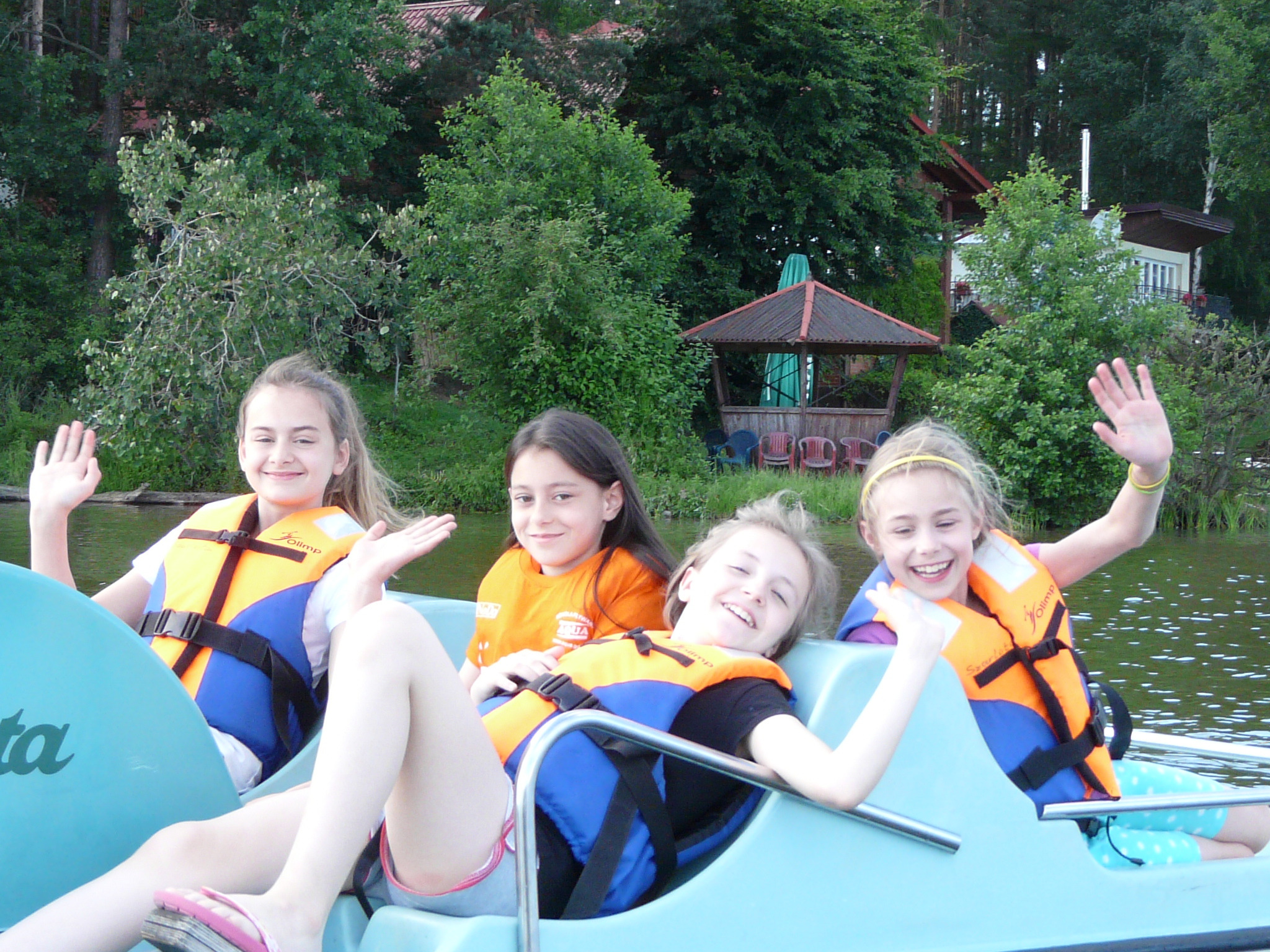 Grupa uśmiechniętych dziewcząt w kamizelkach ratunkowych macha rękami, siedząc na turkusowym rowerze wodnym na jeziorze, w tle widoczny las i altana.