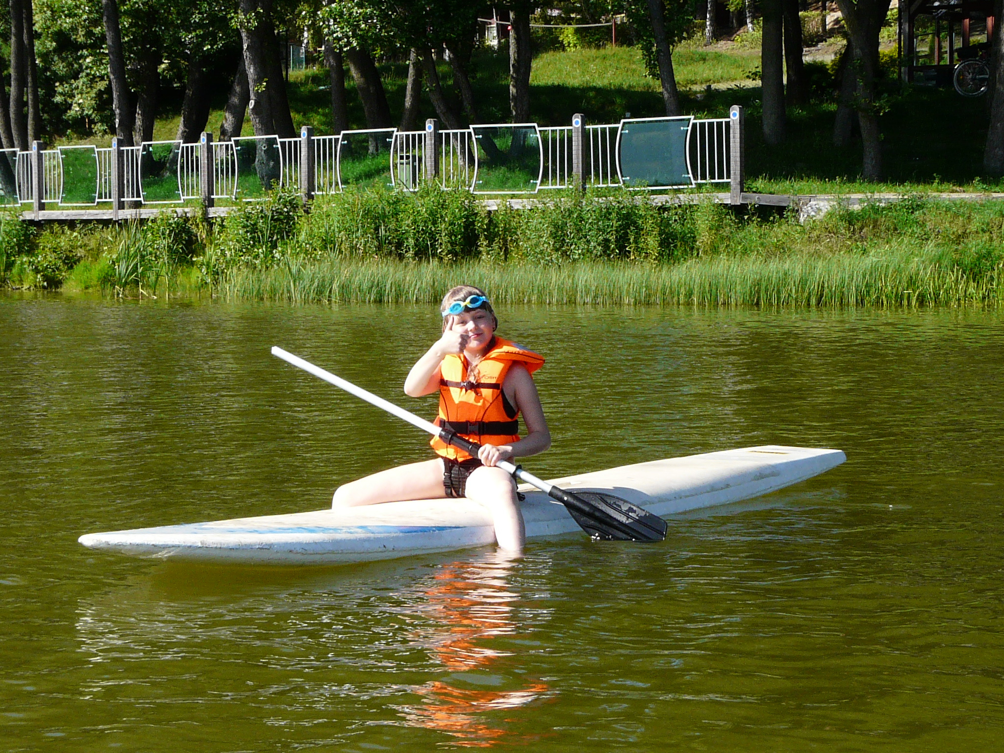 Dziewczyna w pomarańczowej kamizelce asekuracyjnej i niebieskich okularach pływackich siedzi na desce SUP na jeziorze, trzymając wiosło i pokazując kciuk w górę.