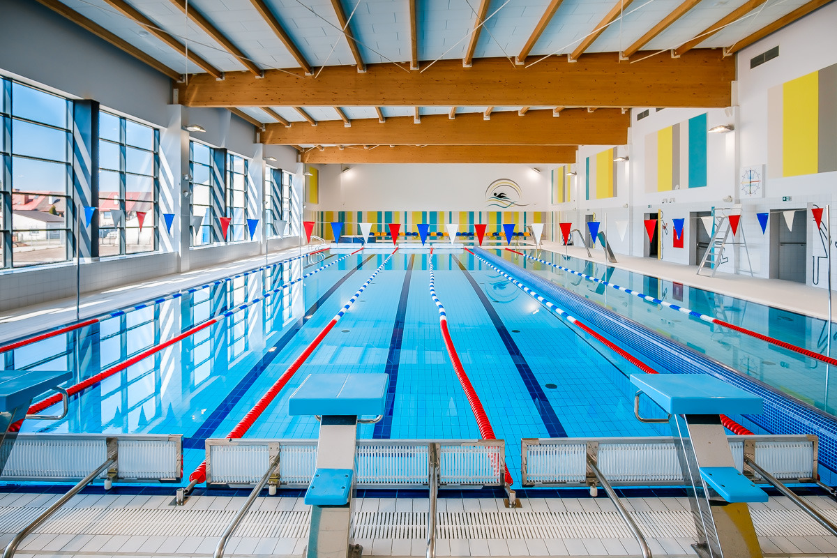 Kryty basen sportowy z niebieską wodą, czerwonymi i białymi linami torów, oraz z niebieskimi słupkami startowymi. W tle widoczne duże okna z widokiem na budynki oraz dekoracyjne, kolorowe panele...