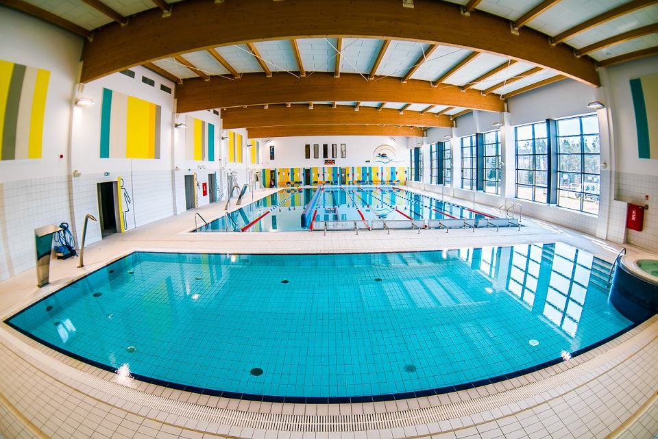 Kryty basen z turkusową wodą, widok z perspektywy rybiego oka, drewniana konstrukcja dachu, duże okna, kolorowe pasy na ścianach.