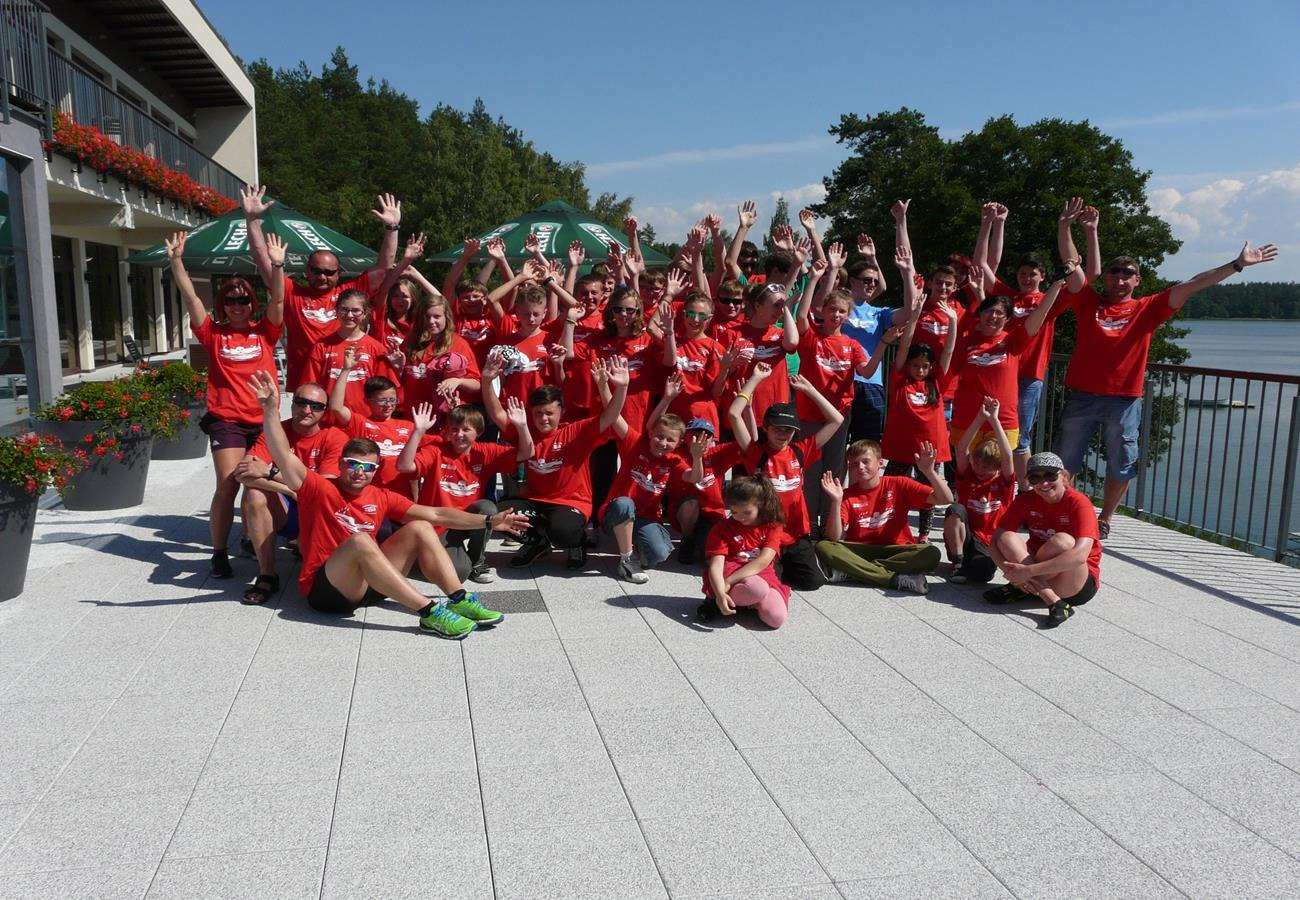 Duża grupa dzieci i dorosłych w czerwonych koszulkach z nadrukiem, z podniesionymi rękami, pozuje na tle budynku z kwiatami i jeziora, prawdopodobnie podczas obozu letniego.