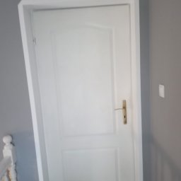 Złota rączka - Białe drzwi wewnętrzne z ościeżnicą po malowaniu, widoczny fragment białej balustrady schodów i szare ściany.