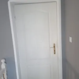 Białe drzwi wewnętrzne z ościeżnicą po malowaniu, widoczny fragment białej balustrady schodów i szare ściany.