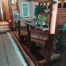 Złota rączka - Drewniana pergola z oświetleniem w Otwocku, widok na taras z deskami, narzędzia i rośliny na stole, wieczorny nastrój.