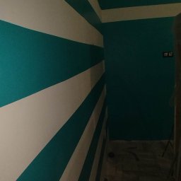 Złota rączka - Ściana w pokoju pomalowana w szerokie, poziome pasy koloru turkusowego i białego, z turkusową ścianą w tle i fragmentem sufitu z pasami, widoczne gniazdka elektryczne.