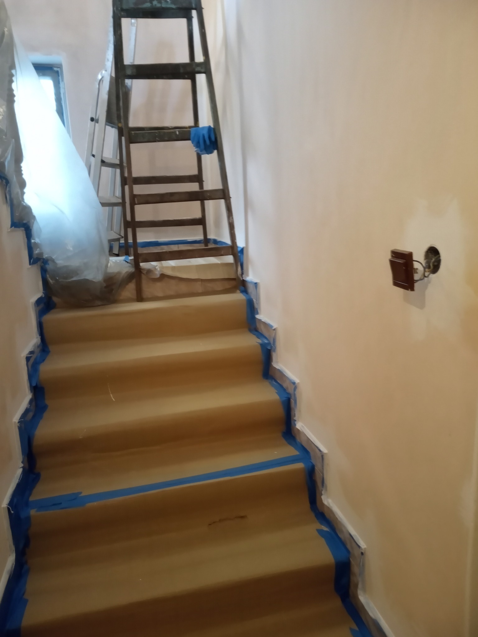 Schody zabezpieczone taśmą malarską i papierem, przygotowane do malowania ścian; drabina oparta o ścianę, w tle okno osłonięte folią malarską.