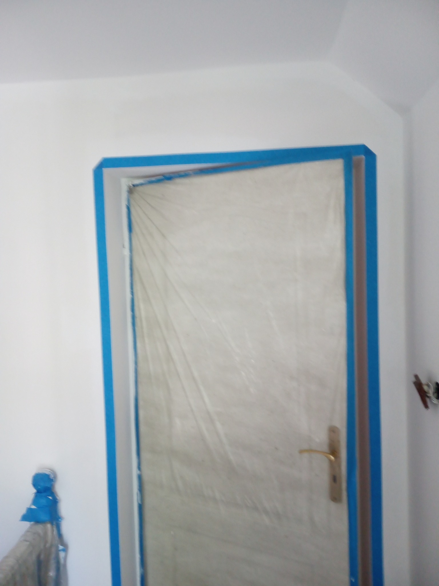 Przygotowanie do malowania: drzwi zabezpieczone folią i taśmą malarską w odcieniach błękitu, ściany w kolorze białym.