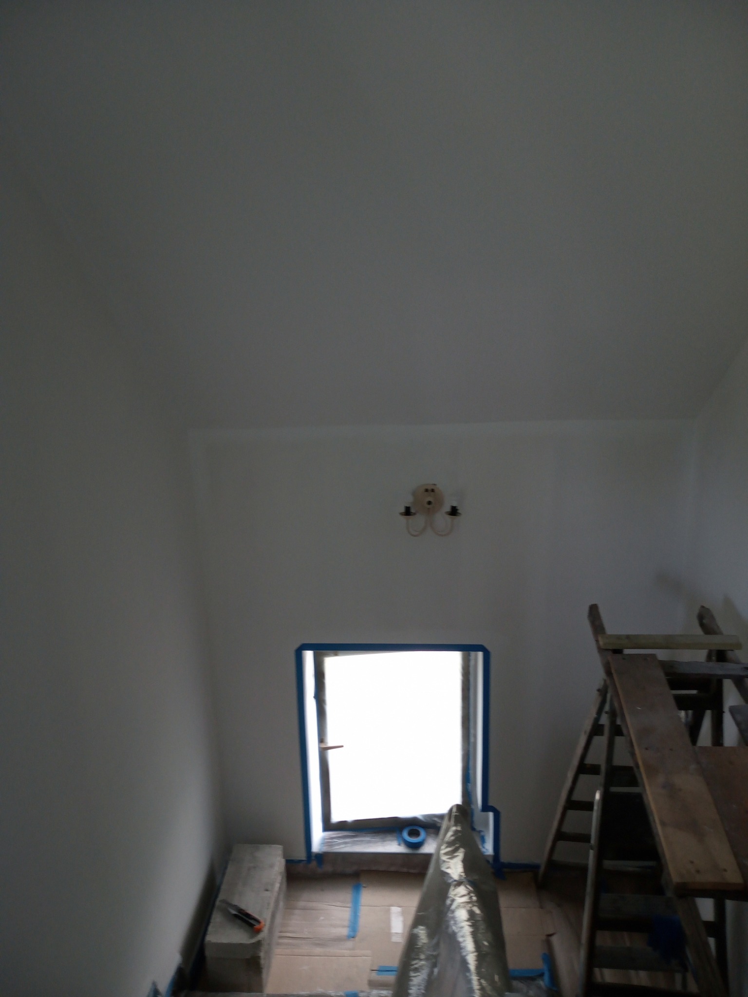 Wnętrze w trakcie odświeżania: ściany pomalowane na biało, okno zabezpieczone taśmą malarską, widoczna drabina i podłoże zabezpieczone folią malarską.