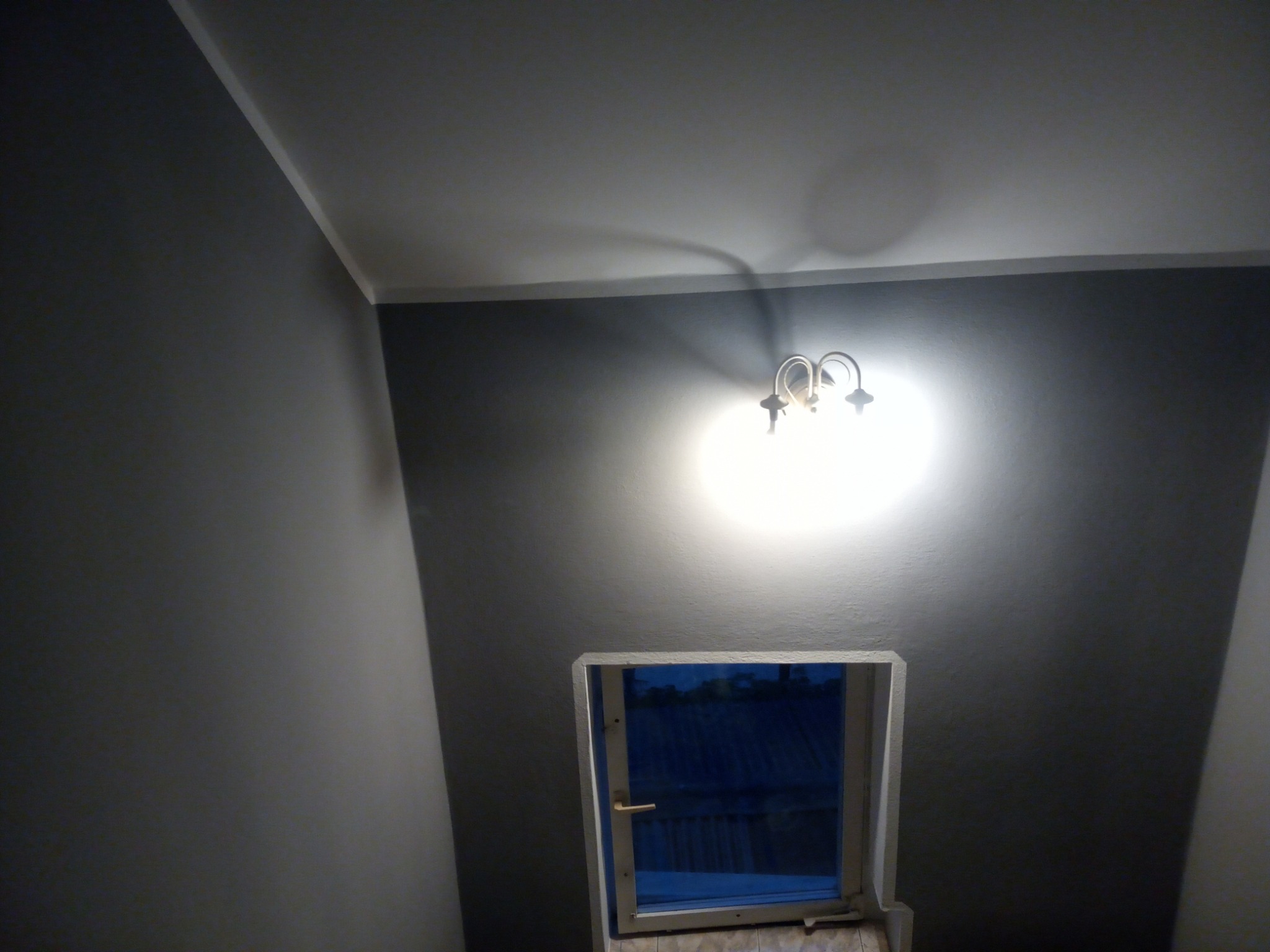 Pokój po odświeżeniu: szare ściany, biały sufit, oświetlenie kinkietem i okno z widokiem na zewnątrz.