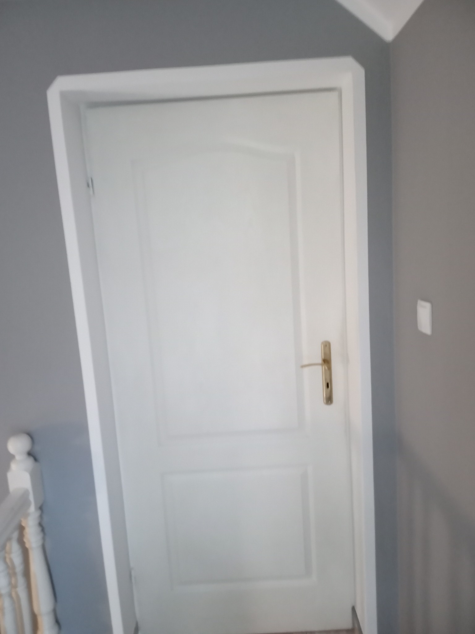 Białe drzwi wewnętrzne z ościeżnicą po malowaniu, widoczny fragment białej balustrady schodów i szare ściany.