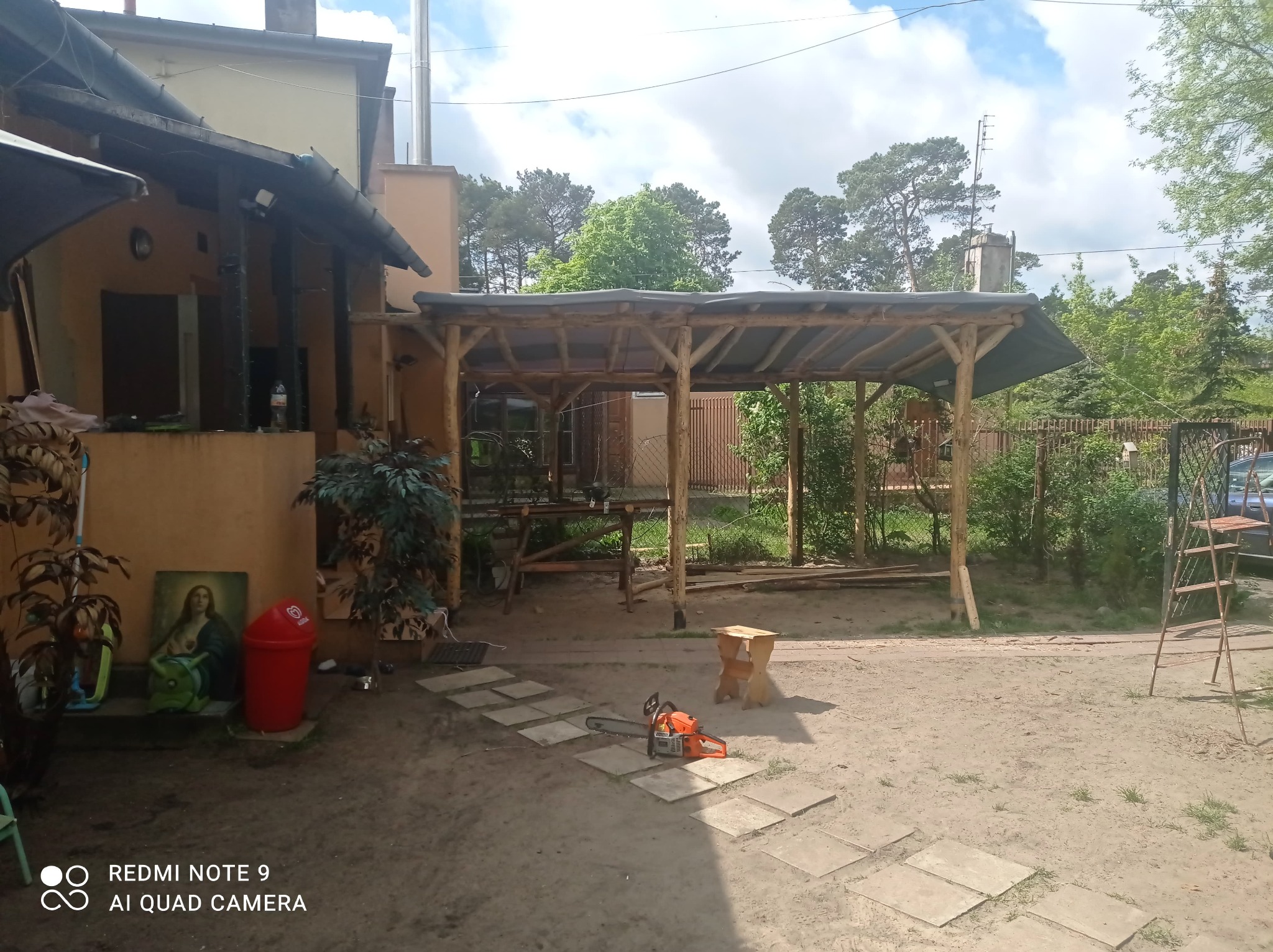 Drewniana pergola z zadaszeniem w trakcie budowy, widoczna piła łańcuchowa leżąca na ścieżce z płyt, drabina oparta o konstrukcję, w tle budynek mieszkalny.