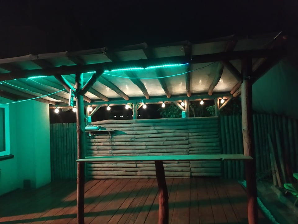 Drewniana pergola z oświetleniem LED i żarówkami, z drewnianym blatem barowym i podłogą z desek, oświetlona nocą.