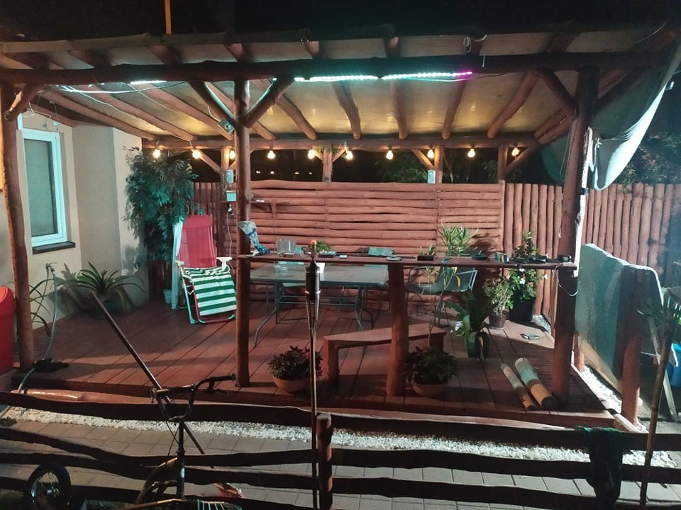 Zadaszony taras z drewnianą pergolą oświetloną girlandą żarówek i taśmą LED, widoczne meble ogrodowe, rośliny w donicach i drewniane ogrodzenie.