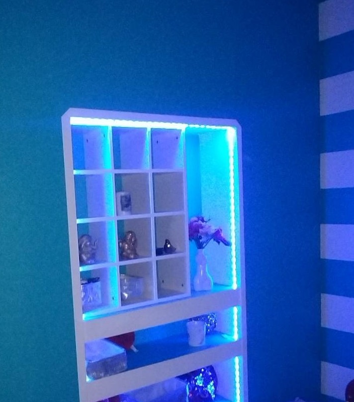 Biały regał z podświetleniem LED, ustawiony na tle turkusowej ściany i ściany w biało-niebieskie pasy, z dekoracyjnymi przedmiotami na półkach.