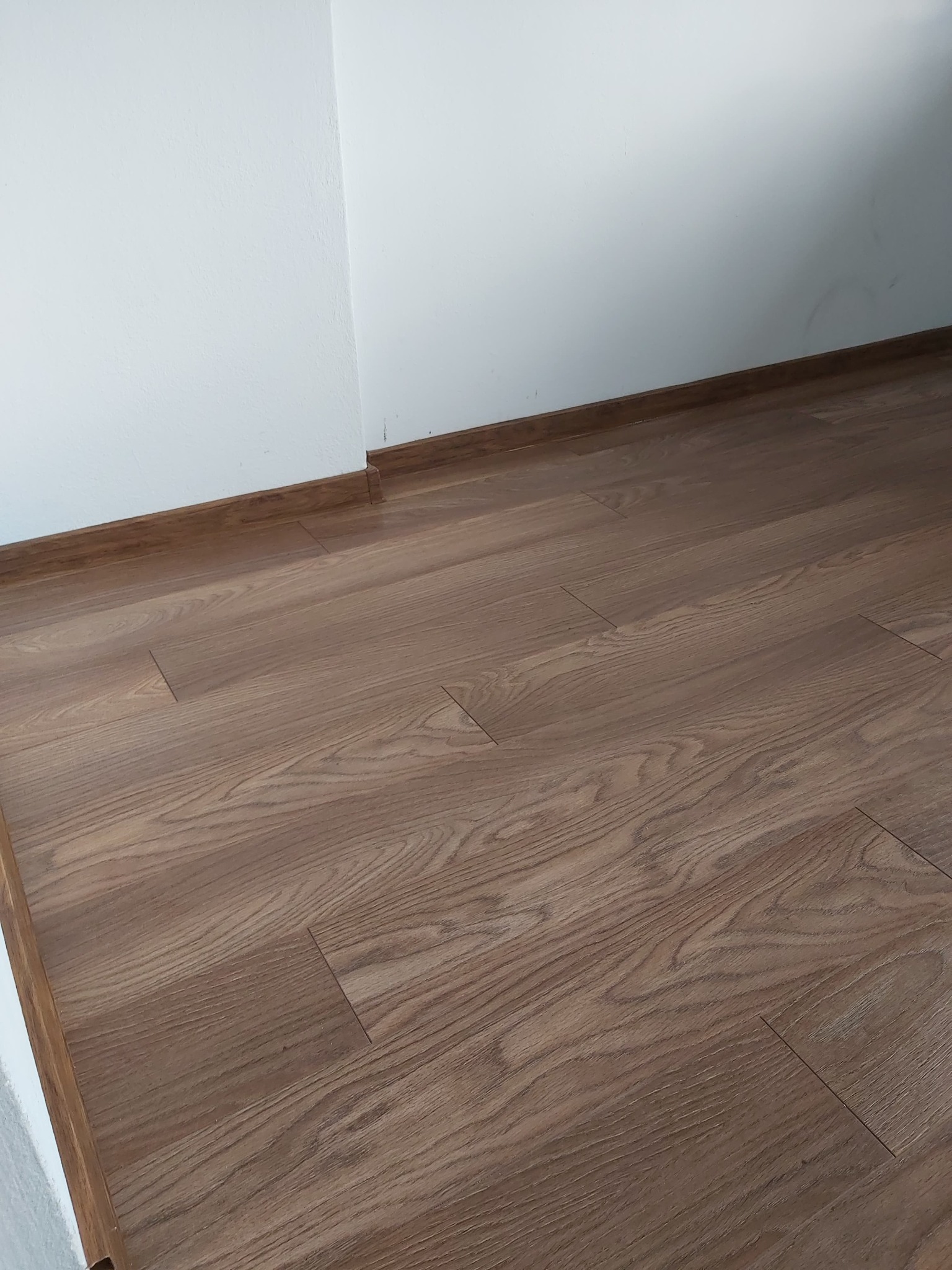 Ułożona podłoga z paneli w odcieniach brązu, widoczna przy białej ścianie z brązową listwą przypodłogową.