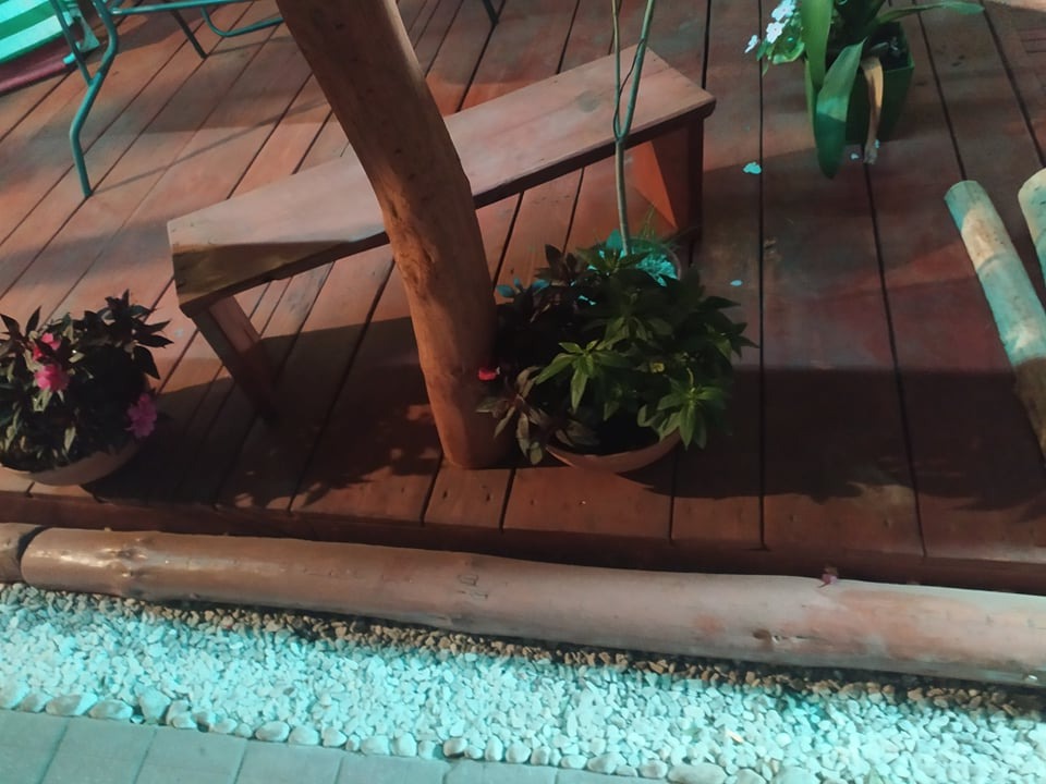 Drewniany taras z donicami kwiatów, ławką z pnia drzewa i obrzeżem z okrąglaków oraz białego grysu.