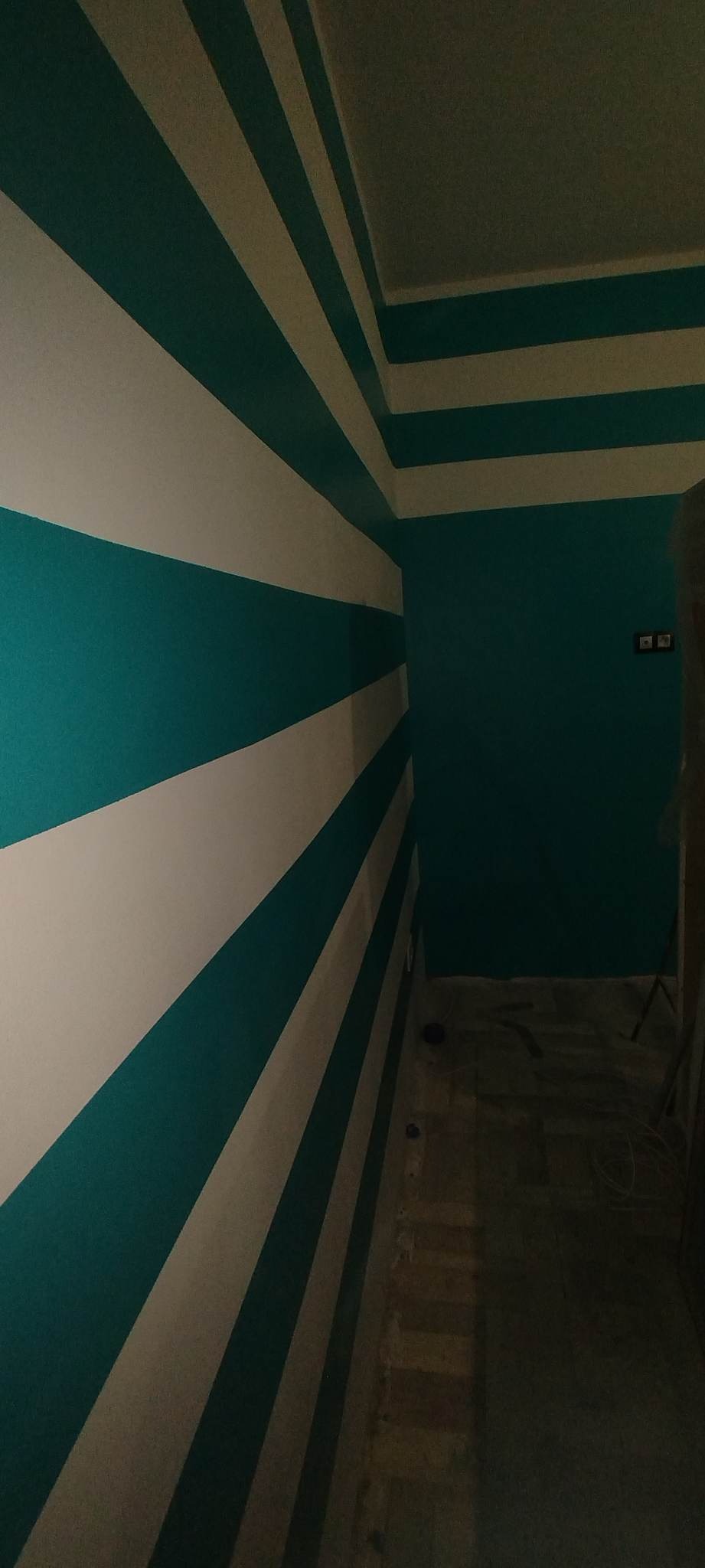 Ściana w pokoju pomalowana w szerokie, poziome pasy koloru turkusowego i białego, z turkusową ścianą w tle i fragmentem sufitu z pasami, widoczne gniazdka elektryczne.