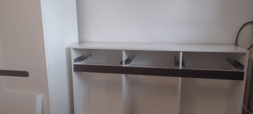 Biała szafka z brązowym akcentem, widoczne prowadnice szuflad, wnętrze szafki puste, surowy stan przed wykończeniem.