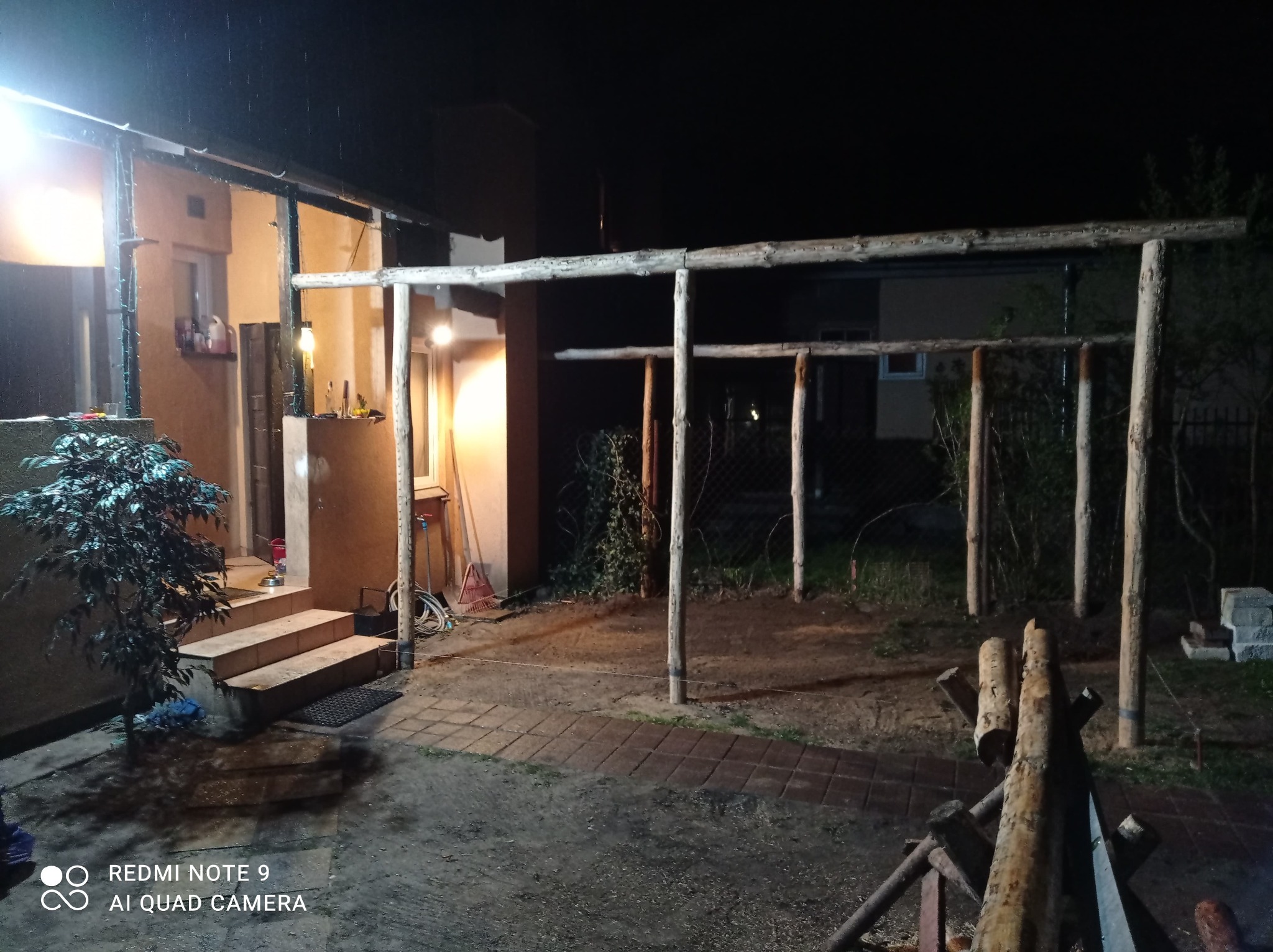 Konstrukcja drewnianej pergoli w trakcie budowy, widoczne surowe drewniane słupy i belki, teren wokół placu budowy częściowo utwardzony kostką, a częściowo ziemny, wykonane w nocy.