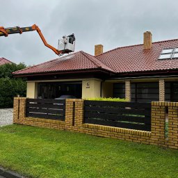 Twoje4sciany - Pracownik na podnośniku koszowym czyści dach domu z czerwonej dachówki za pomocą myjki ciśnieniowej, widoczny fragment żółto-ceglanego ogrodzenia i trawnik.