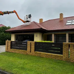 Pracownik na podnośniku koszowym czyści dach domu z czerwonej dachówki za pomocą myjki ciśnieniowej, widoczny fragment żółto-ceglanego ogrodzenia i trawnik.