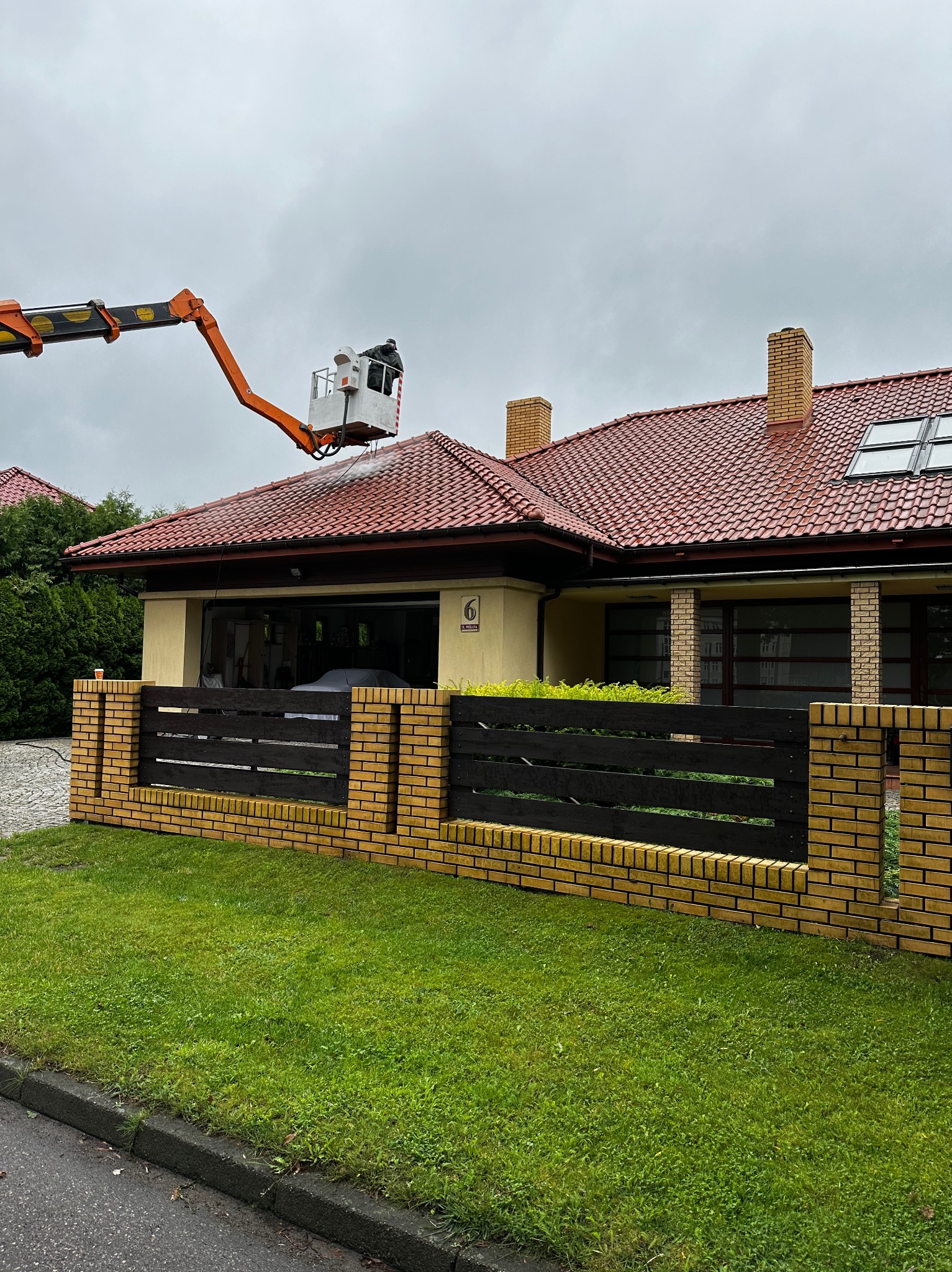Pracownik na podnośniku koszowym czyści dach domu z czerwonej dachówki za pomocą myjki ciśnieniowej, widoczny fragment żółto-ceglanego ogrodzenia i trawnik.