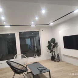 Nowoczesny salon z oświetleniem szynowym, minimalistycznym stolikiem kawowym, plecionymi krzesłami i telewizorem na białej ścianie, podkreślony przez roślinę doniczkową i dekoracyjne lampiony.