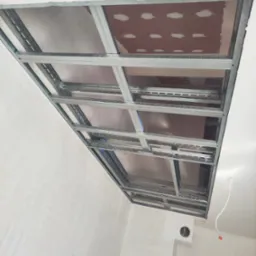 Szkielet metalowy sufitu podwieszanego z widocznymi płytami w kolorze brązowym, częściowo zamontowany, z perspektywy dolnej, widoczne kable elektryczne i fragment ściany.