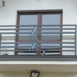 Szara, metalowa balustrada balkonowa z poziomymi prętami i geometrycznym wzorem na tle okna i białej ściany budynku.