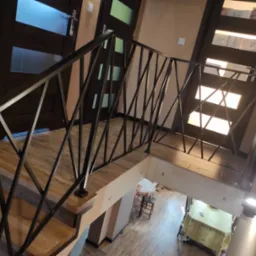 Metalowa balustrada schodowa z geometrycznym wzorem, widok z góry na drewniane stopnie i fragment pomieszczenia poniżej.