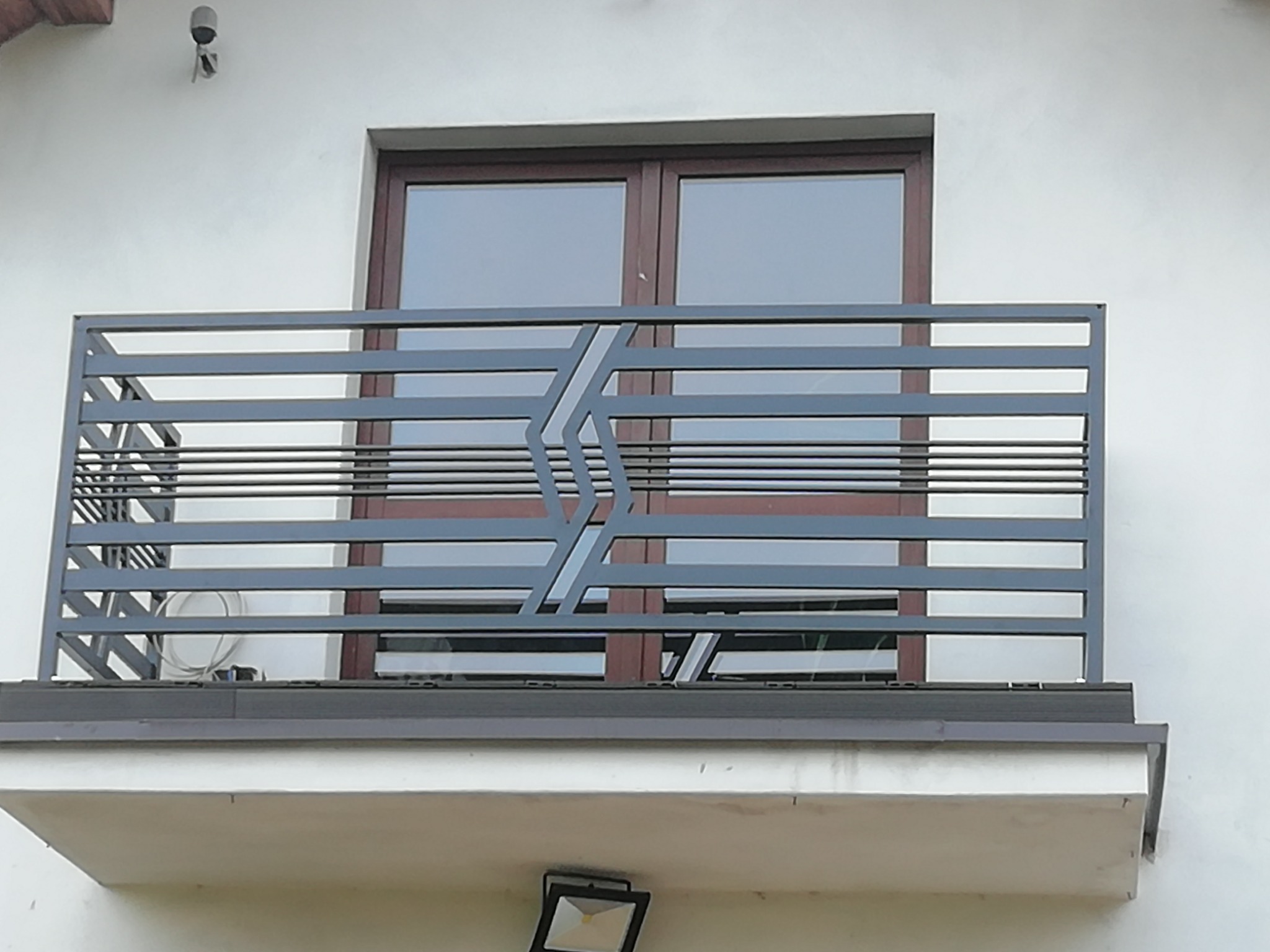 Szara, metalowa balustrada balkonowa z poziomymi prętami i geometrycznym wzorem na tle okna i białej ściany budynku.