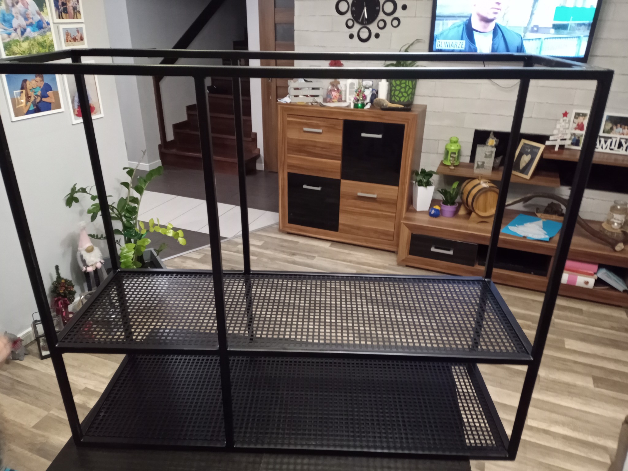 Czarna, metalowa konstrukcja regału z dwoma półkami z siatki, na tle wnętrza domu z drewnianymi schodami i meblami.