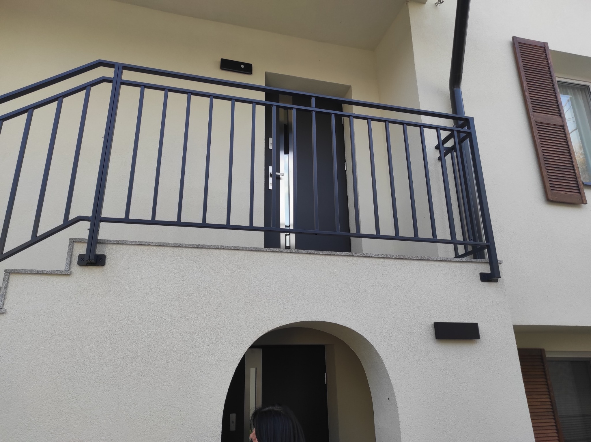Czarne, metalowe balustrady schodowe i balkonowe z pionowymi szczeblami, zamontowane na jasnym budynku z łukowatym wejściem i brązowymi okiennicami