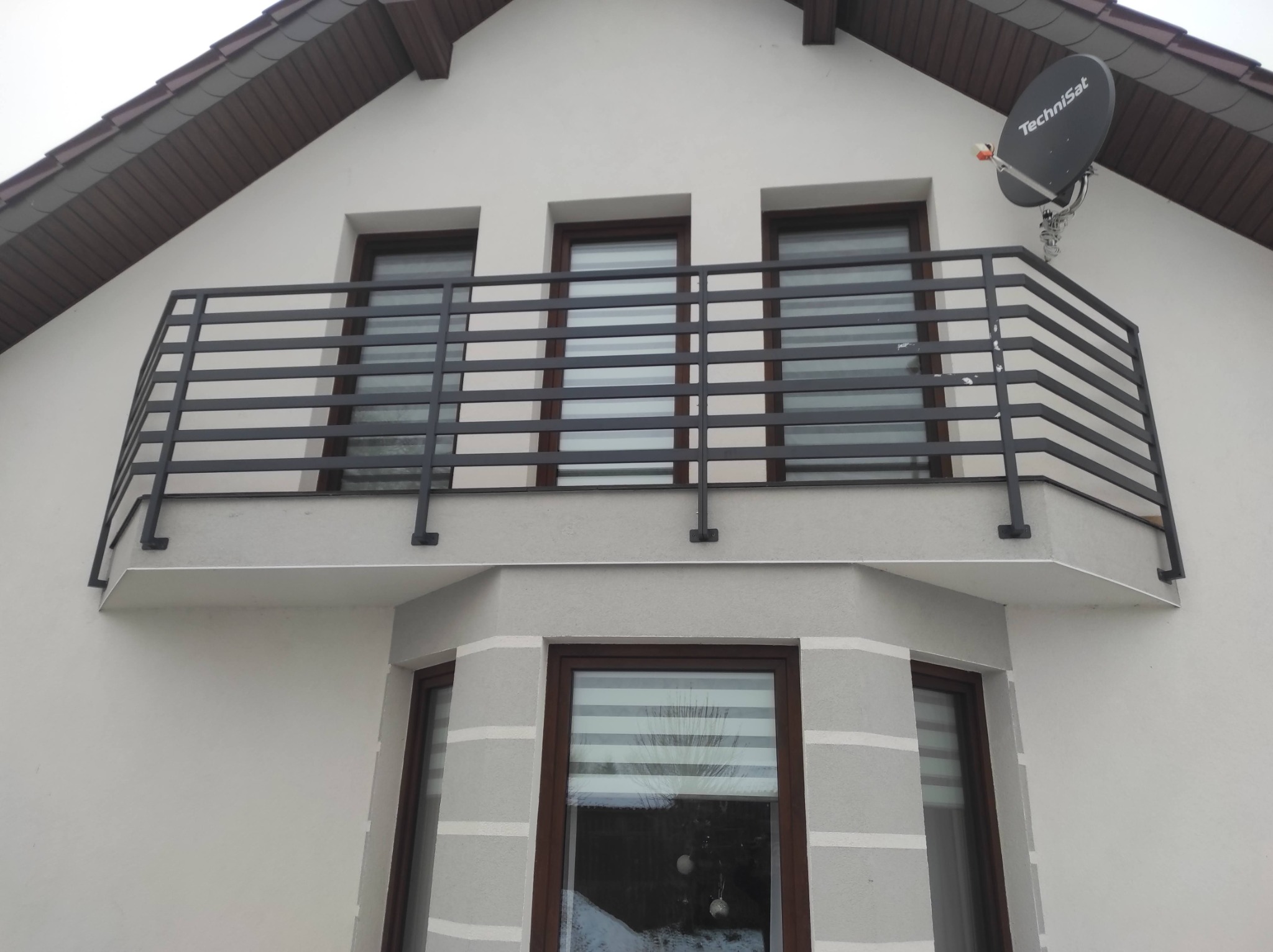 Czarna, pozioma balustrada balkonowa z prostokątnych profili, zamontowana na balkonie domu jednorodzinnego; widoczny fragment elewacji z trzema oknami i anteną satelitarną.