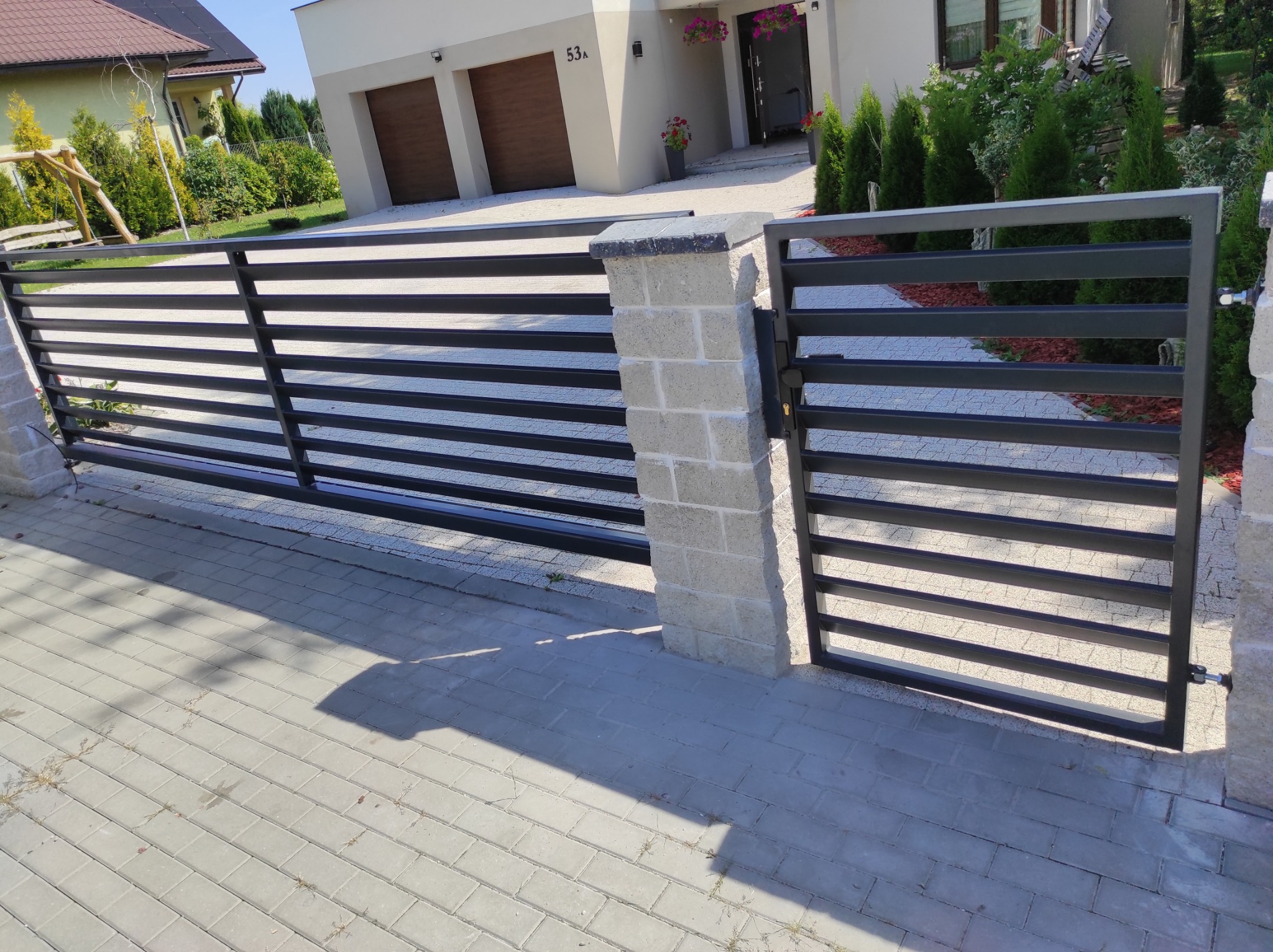 Nowoczesna, pozioma brama wjazdowa z czarnych profili stalowych, osadzona na murowanych słupkach z szarej kostki, na tle domu z garażem i zadbanego ogrodu.