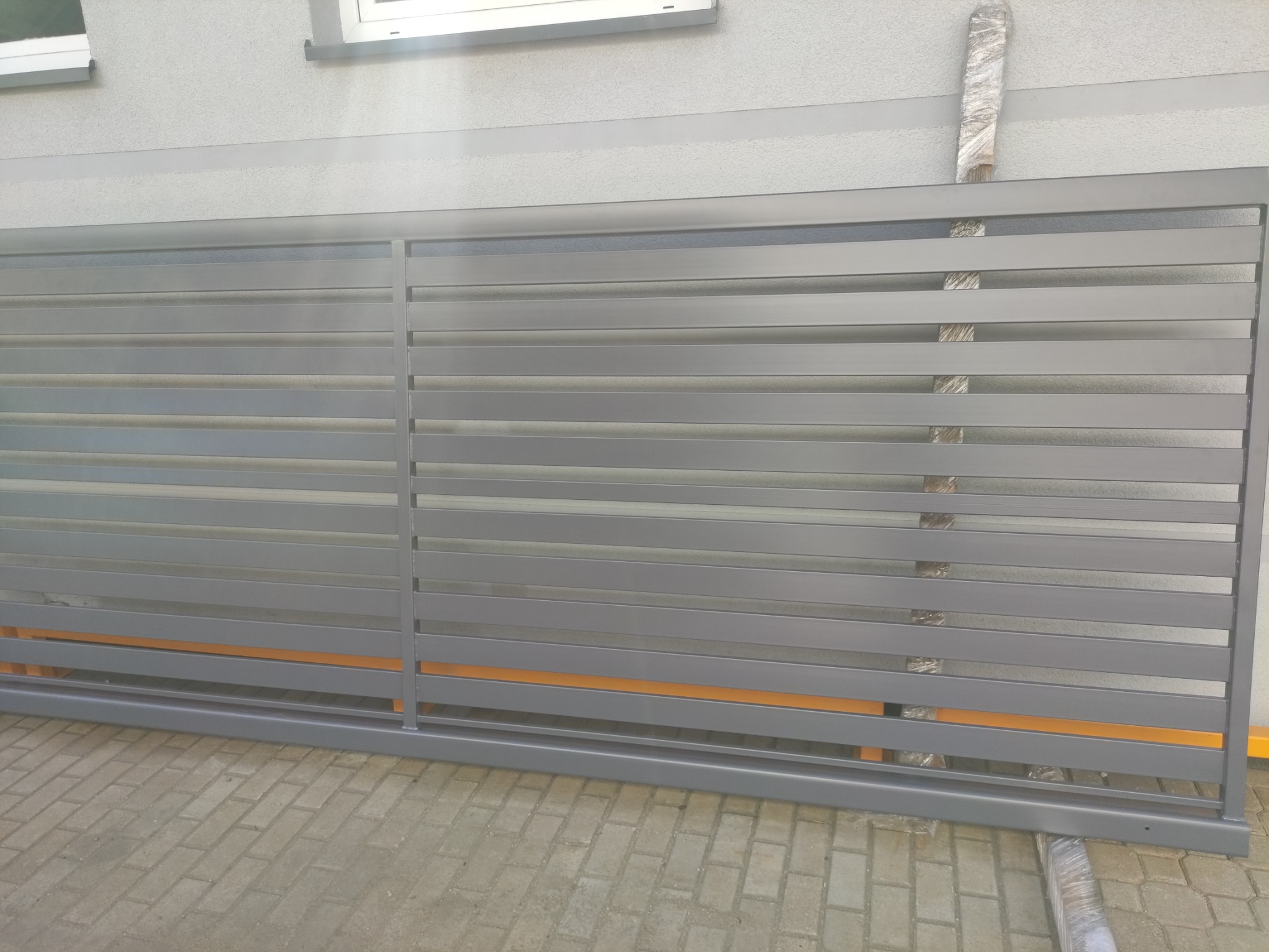 Nowoczesna przesuwna brama wjazdowa z poziomych, szarych profili, częściowo zabezpieczona folią ochronną, na tle jasnoszarej elewacji.