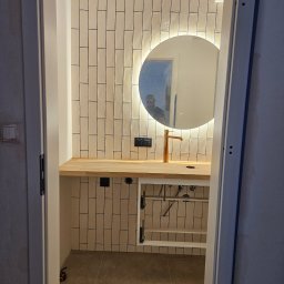 ZAZA NATCHKEBIA GMP-BUD - Wykończona łazienka z beżowymi, pionowymi płytkami, drewnianym blatem, okrągłym lustrem z podświetleniem LED i złotą baterią. Widok zza otwartych drzwi.