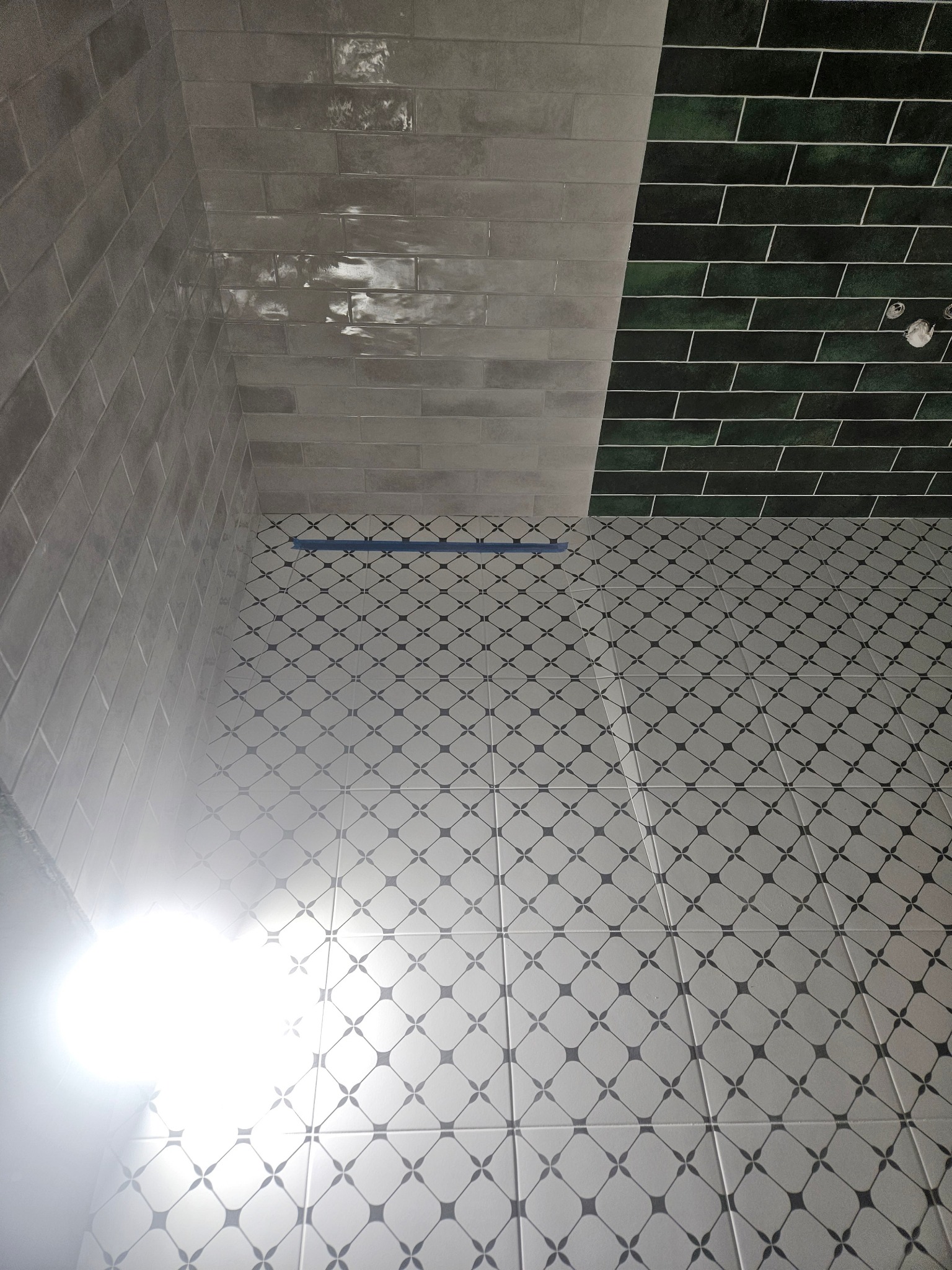 Ukończona łazienka z mozaikową podłogą, szarymi, błyszczącymi płytkami ściennymi ułożonymi w cegiełkę oraz ciemnozielonymi płytkami w górnej części ściany, oddzielonymi niebieską taśmą malarską.