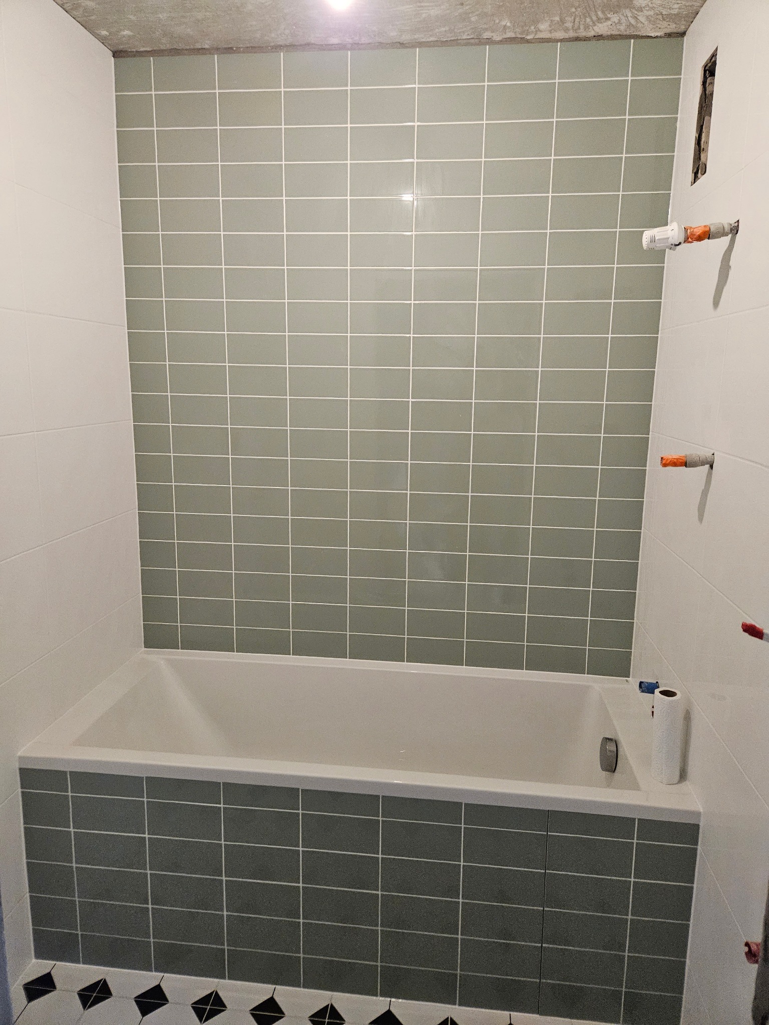 Łazienka w trakcie remontu: ściany wyłożone szarymi płytkami z białą fugą, zabudowana wanna, widoczne niedokończone instalacje i surowy betonowy sufit.