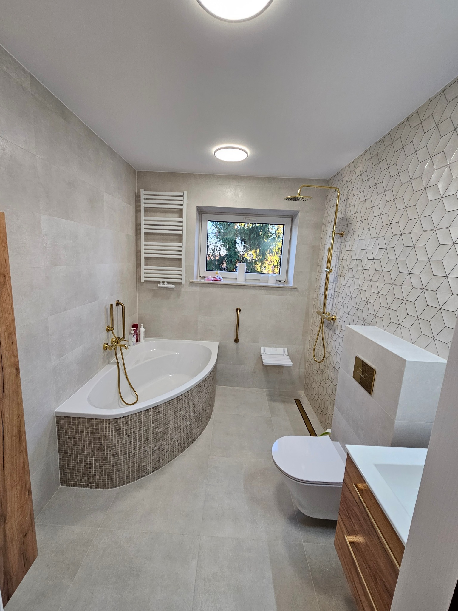Nowoczesna łazienka z narożną wanną obudowaną mozaiką, złotą armaturą i geometryczną płytką na ścianie, widok na okno.