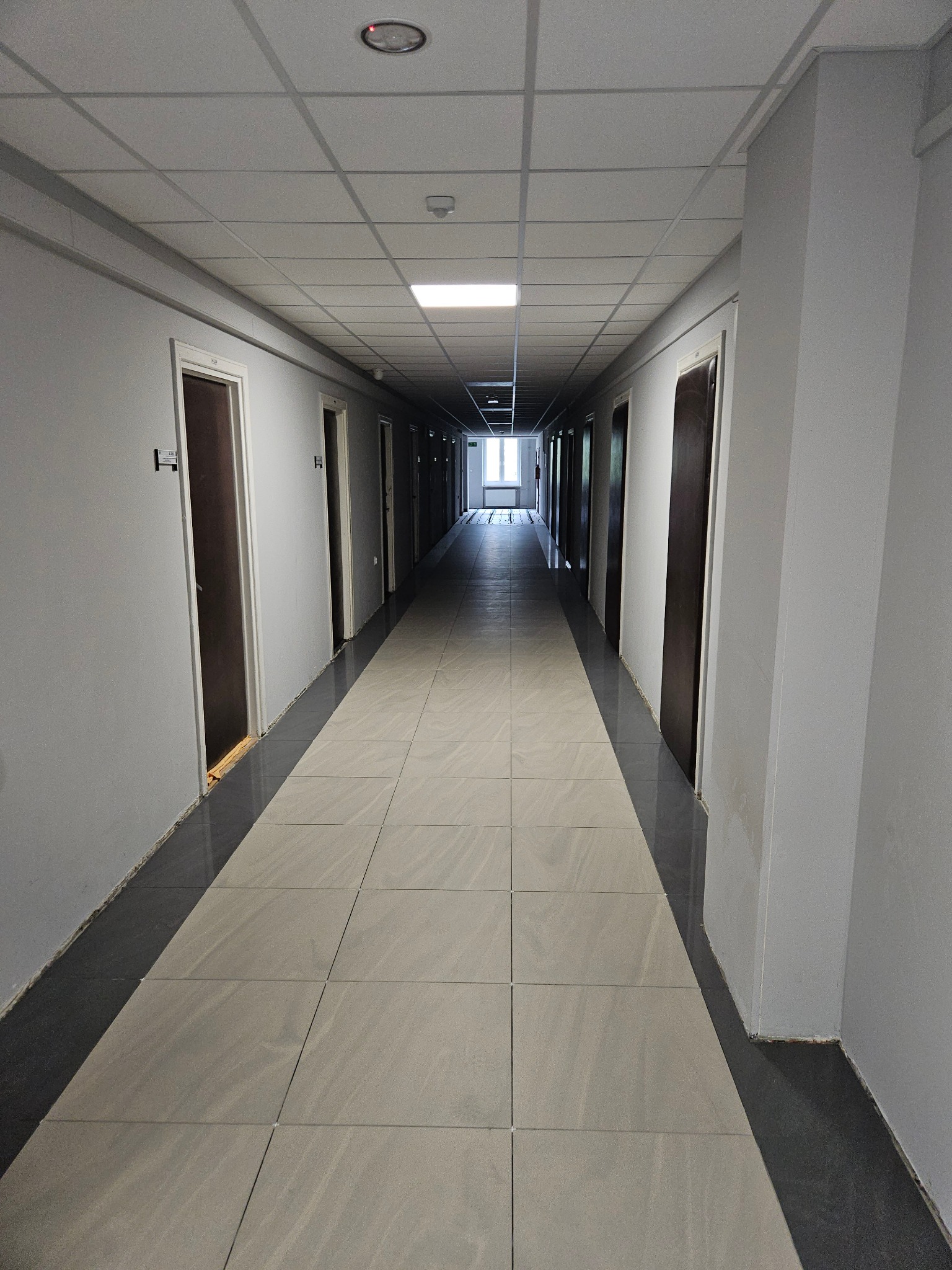 Długi korytarz w budynku biurowym z szarymi ścianami, ciemnymi drzwiami i płytkami podłogowymi w odcieniach szarości, widoczne niedociągnięcia przy listwach podłogowych.