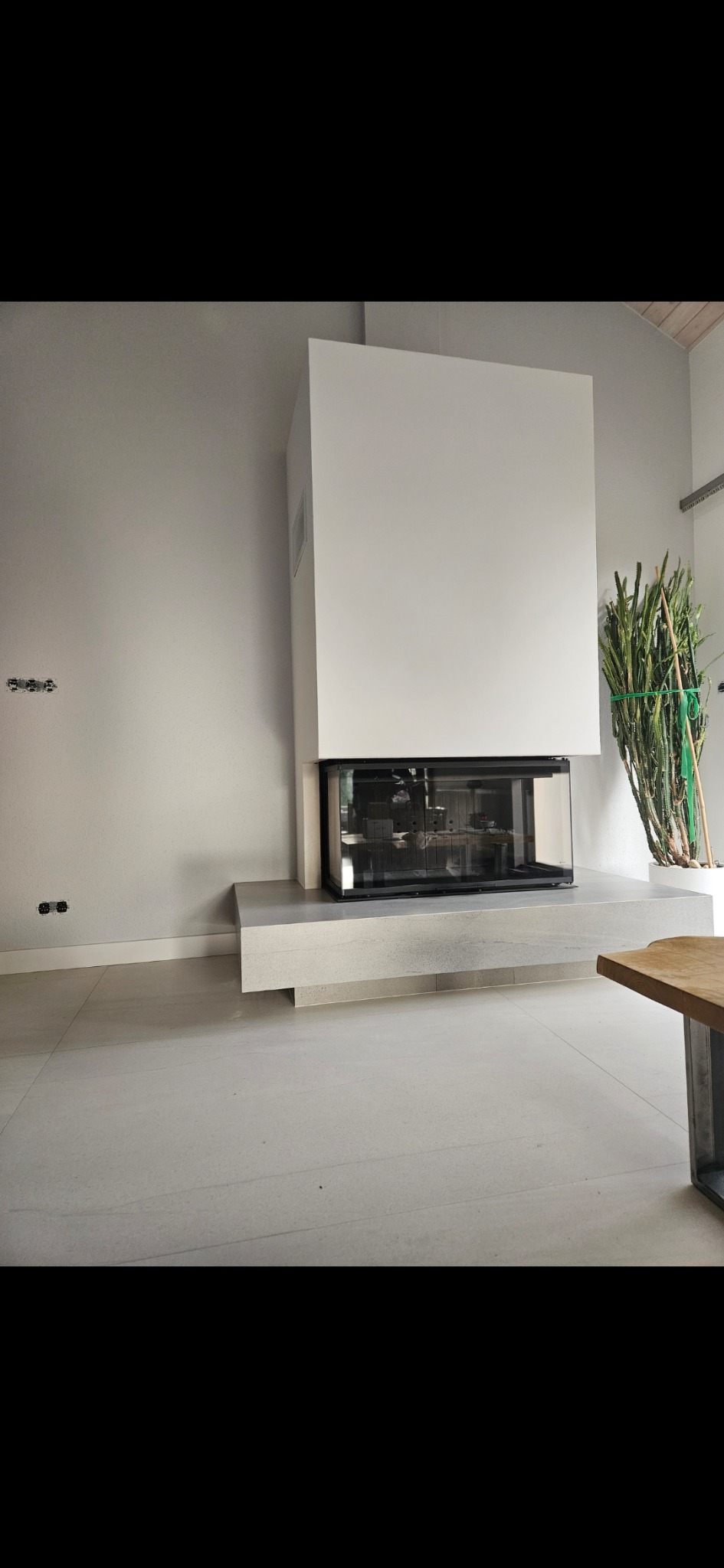 Nowoczesny kominek z przeszklonym paleniskiem, obudowany białym blokiem, na tle jasnej ściany i podłogi. Minimalistyczny design wnętrza z rośliną w tle.