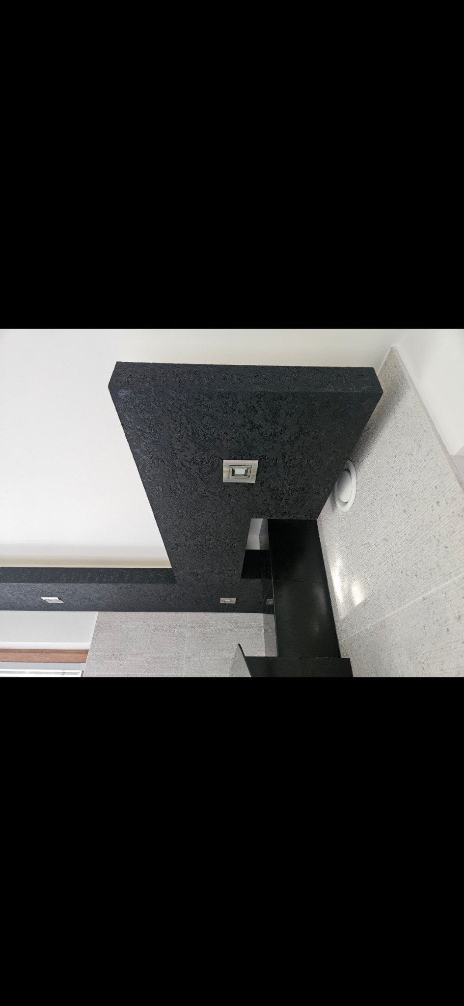 Nowoczesny sufit podwieszany z czarną, teksturowaną płytą i wbudowanym oświetleniem LED, kontrastujący z jasnymi płytkami ściennymi w łazience. Widoczny fragment wentylacji.