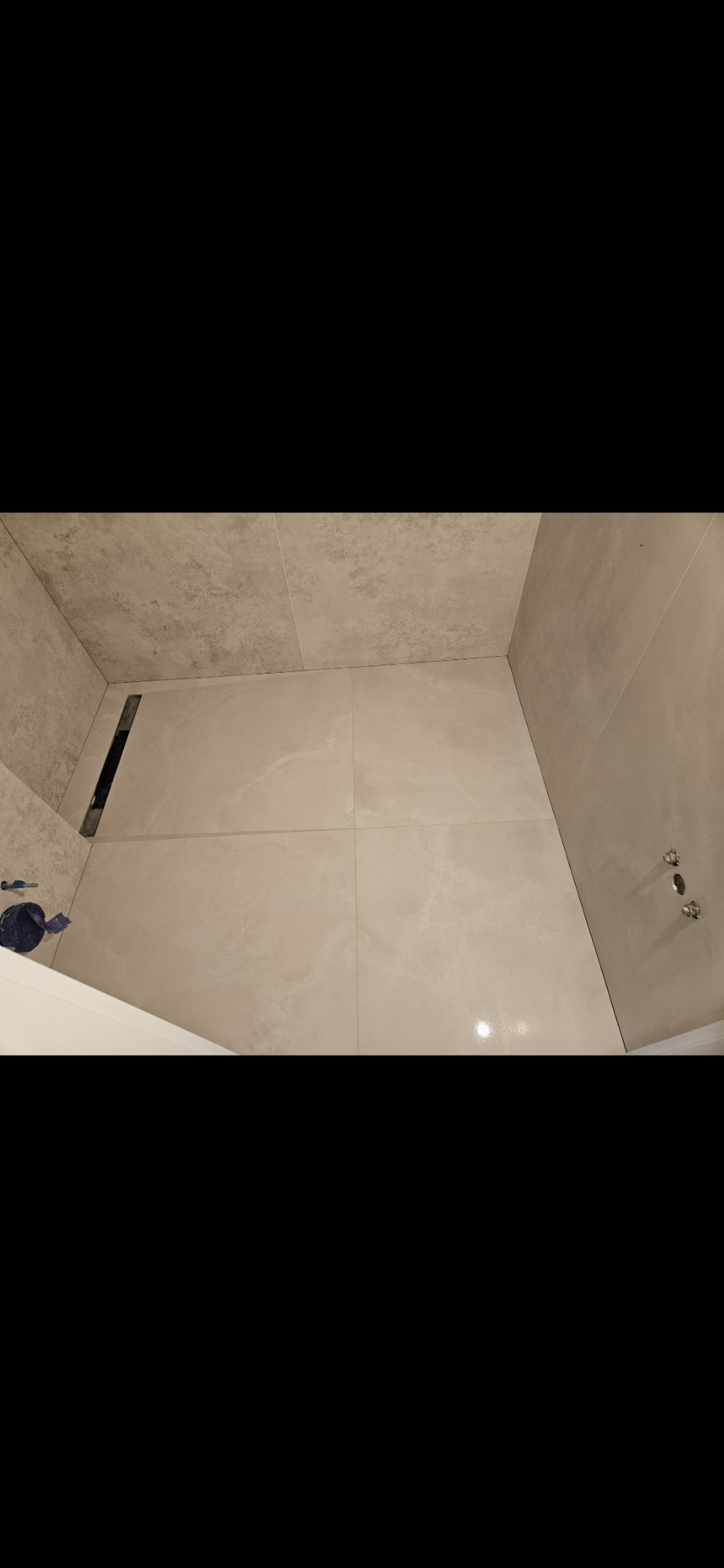 Wykończona łazienka z beżowymi płytkami, odpływem liniowym i wystającymi rurami. Widok z góry na podłogę i fragment ściany. Minimalistyczny design.