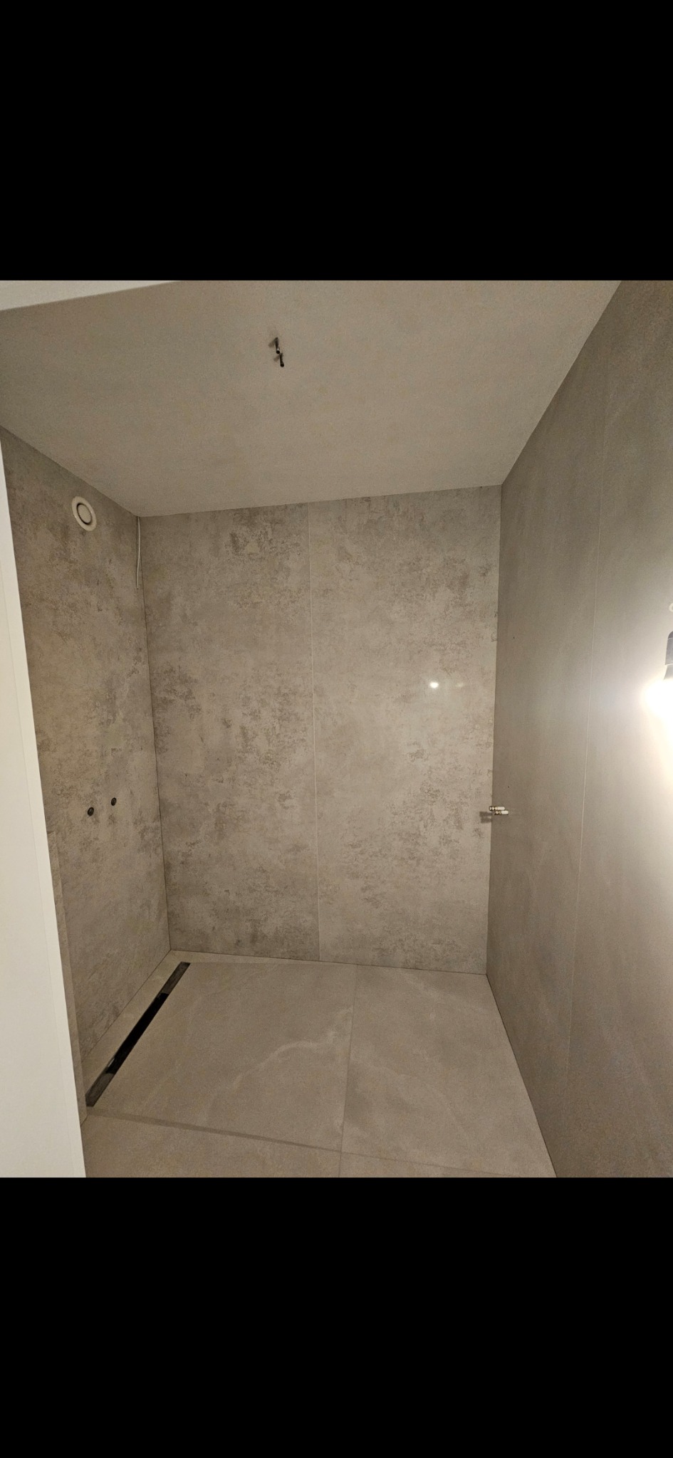 Wykończona kabina prysznicowa z odpływem liniowym i szarymi płytkami imitującymi kamień. Widoczne punkty podłączenia armatury. Minimalistyczny design.