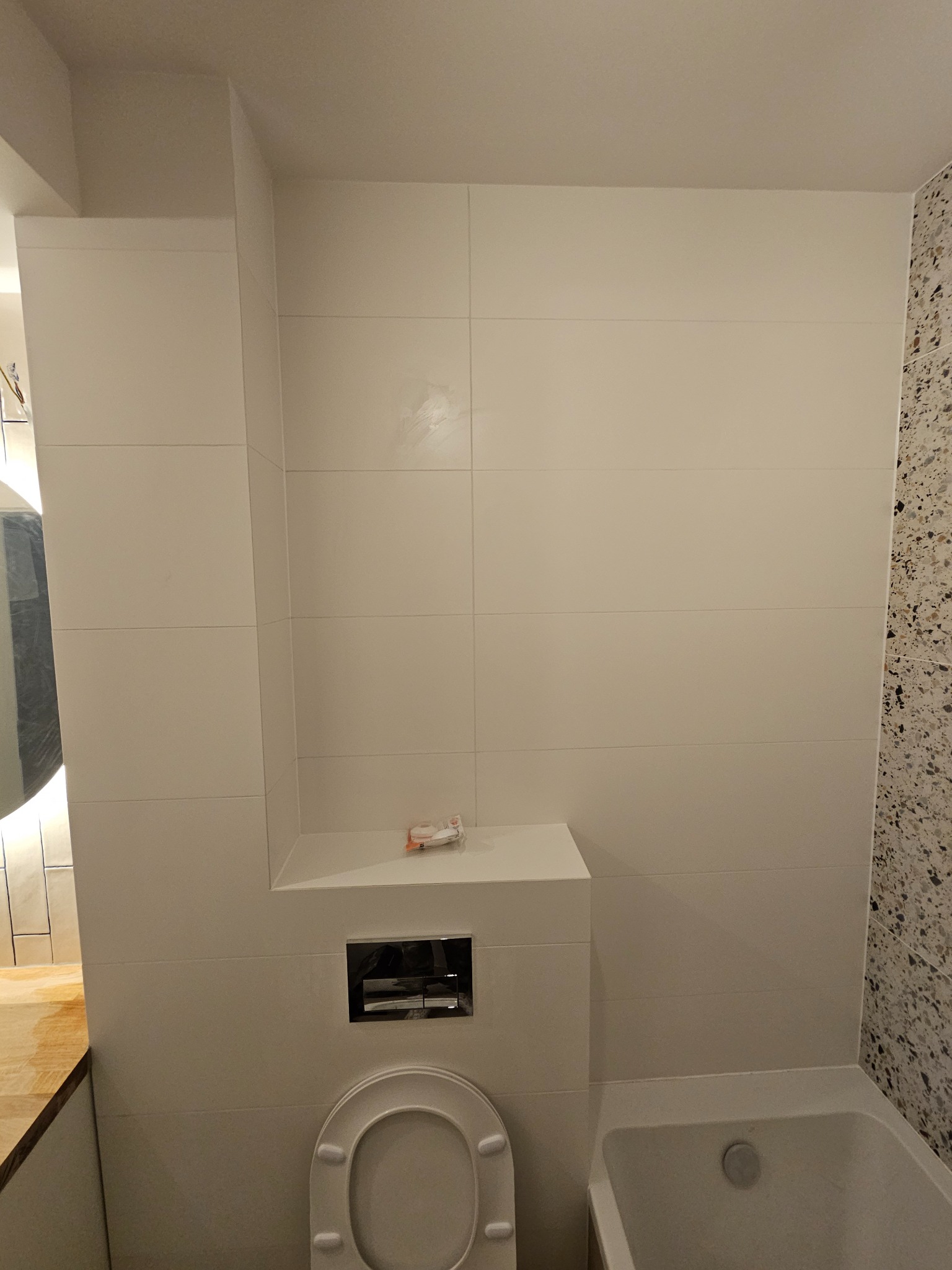 Biała łazienka po remoncie: płytki ścienne, zabudowa WC z przyciskiem spłukującym, fragment wanny i mozaika na ścianie. Minimalistyczny design.