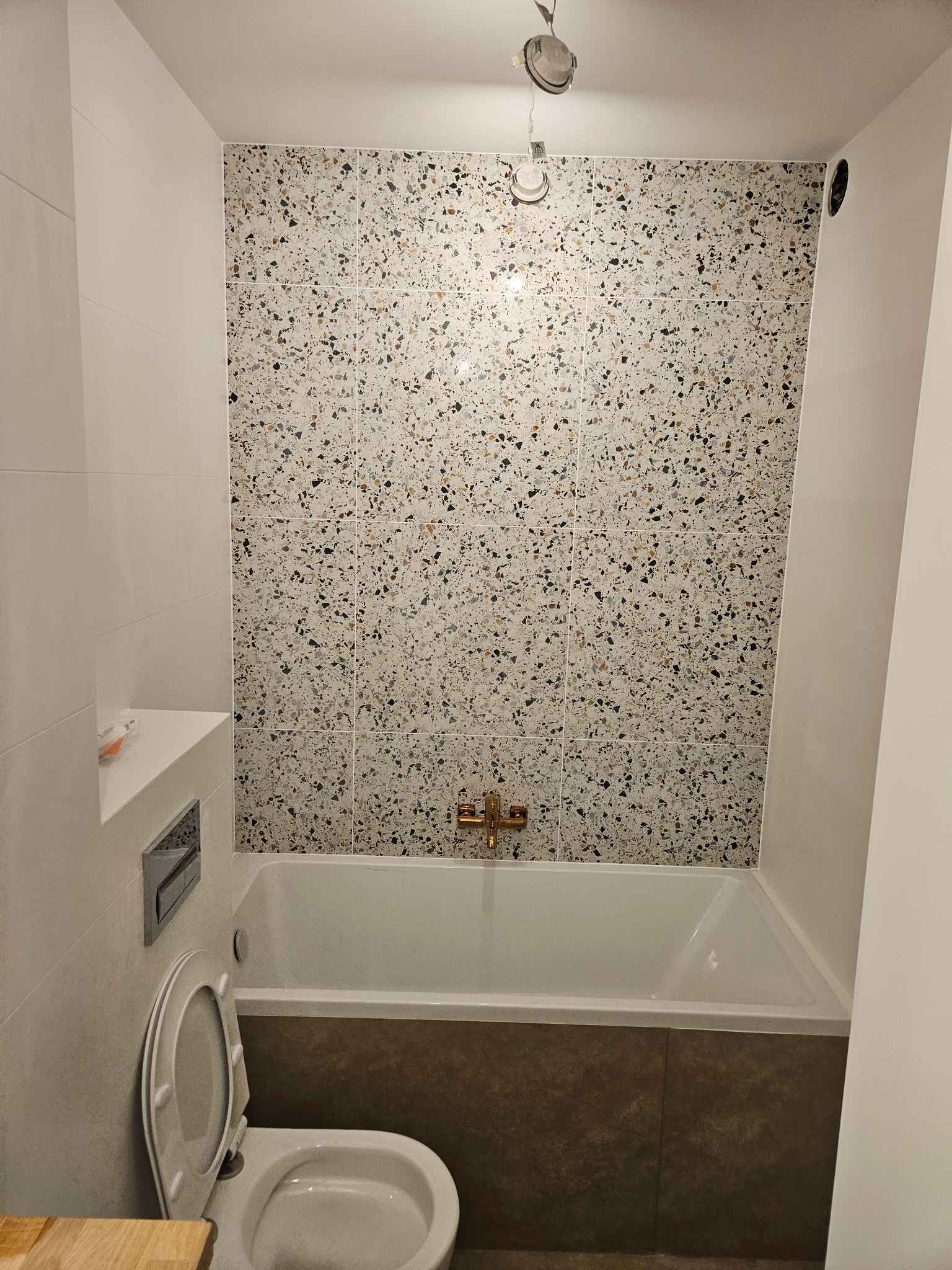 Wykończona łazienka z wanną obudowaną brązowymi płytkami i ścianą w mozaikę. Widoczny fragment toalety i wnęka w ścianie. Oświetlenie sufitowe.