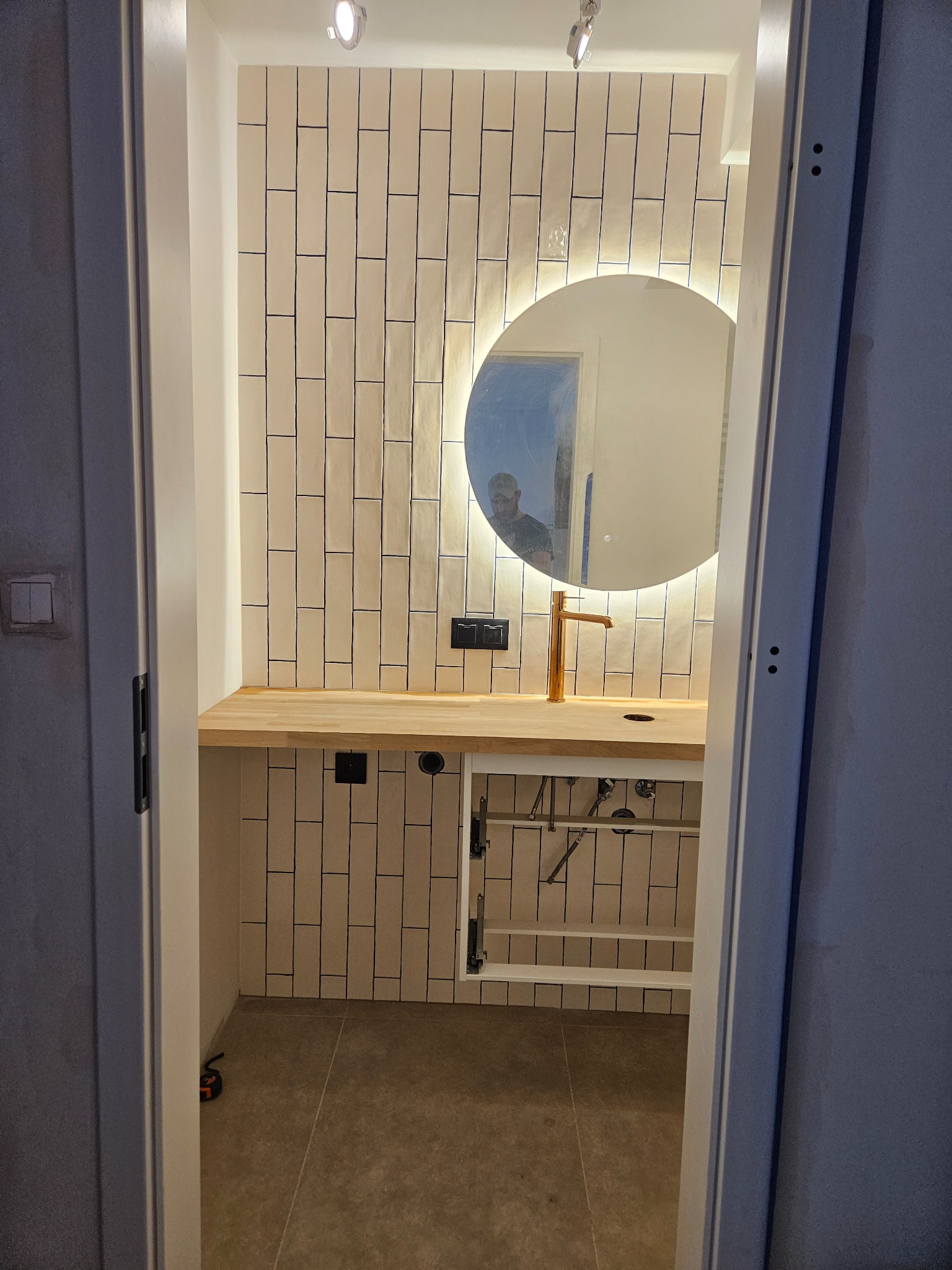 Wykończona łazienka z beżowymi, pionowymi płytkami, drewnianym blatem, okrągłym lustrem z podświetleniem LED i złotą baterią. Widok zza otwartych drzwi.